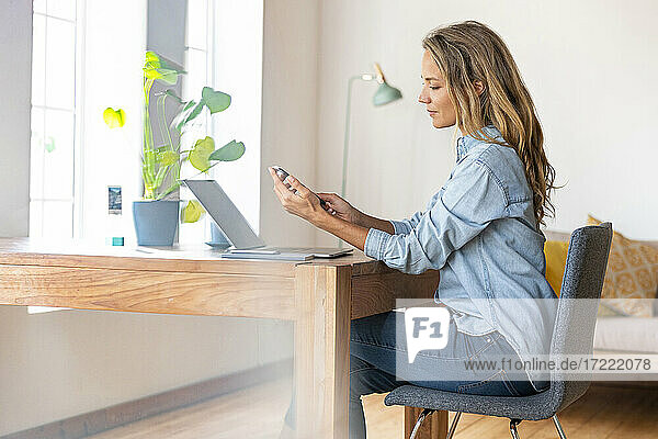 Berufstätige Frau  die im Büro zu Hause ein Mobiltelefon über einem Laptop benutzt