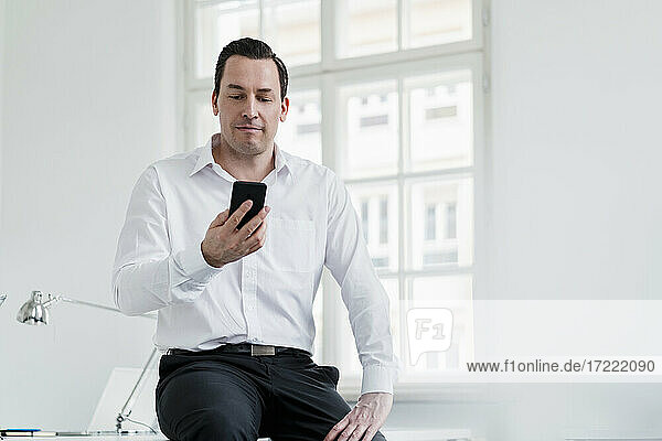 Männlicher Berufstätiger  der am Arbeitsplatz sitzend ein Smartphone benutzt
