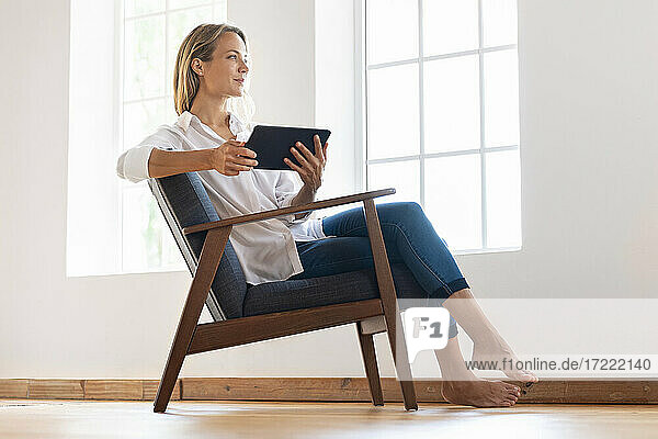 Contemplating woman holding digital tablet while sitting on armchair at home