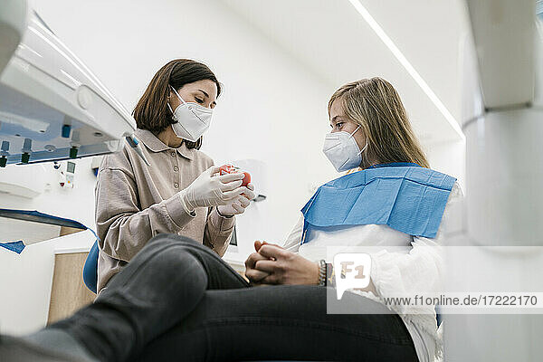 Weiblicher Kieferorthopäde mit Gesichtsmaske  der einem Patienten in der Praxis ein Modell künstlicher Zähne erklärt