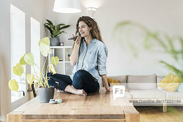 Smiling woman with crossed legs talking on mobile phone at home