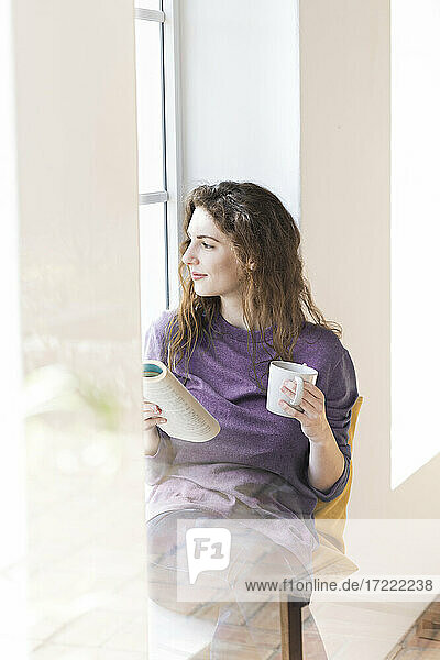 Frau träumt  während sie eine Kaffeetasse und ein Buch am Fenster hält