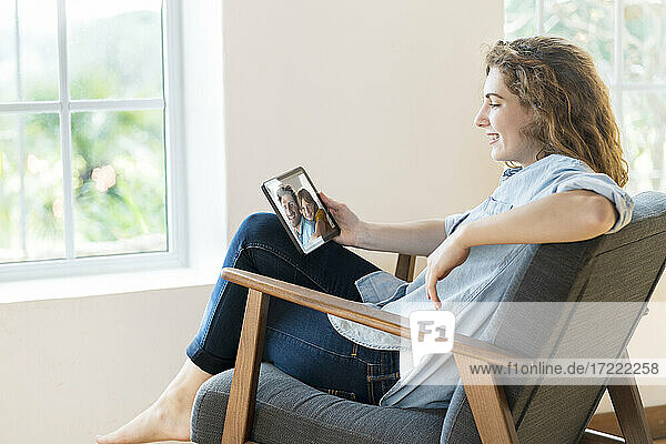 Lächelnde entspannte Frau  die auf einem Sessel im Wohnzimmer sitzt und über ein digitales Tablet einen Videoanruf führt