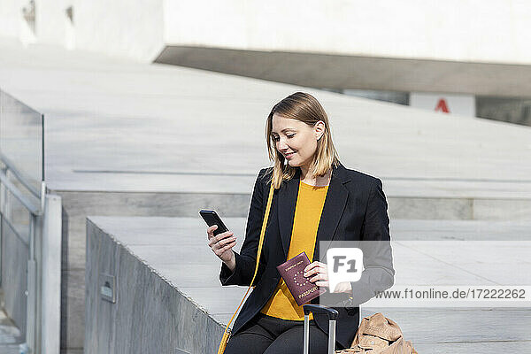 Unternehmer mit Reisepass  der ein Mobiltelefon benutzt  während er auf einer Stützmauer sitzt