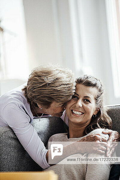 Liebevolle Großmutter  die eine lächelnde Frau umarmt  während sie zu Hause auf dem Sofa sitzt