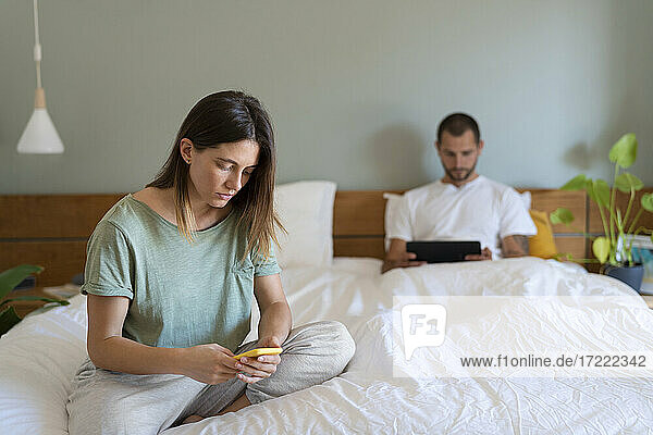 Junge Frau  die auf dem Bett ihr Smartphone benutzt  während ihr Freund im Hintergrund ein digitales Tablet betrachtet