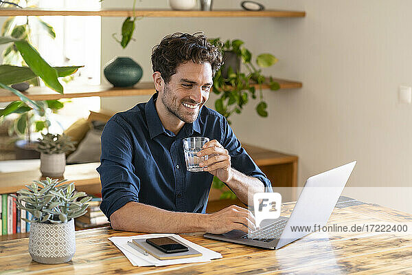 Smiling male freelance worker holding glass of water while working on laptop at home office