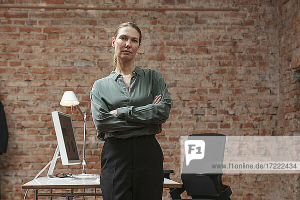 Confident businesswoman with arms crossed standing against desk in office