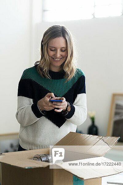 Mittlere erwachsene Frau lächelt  während sie ein Foto von einer Schachtel mit dem Handy zu Hause macht