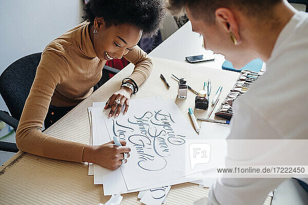 Lächelnde afrikanische Frau schreibt Kalligraphie auf Papier  während sie mit ihrem Freund im Atelier sitzt