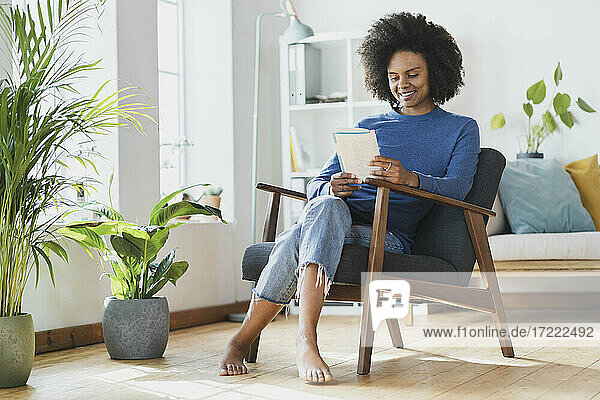Lächelnde Frau liest ein Buch  während sie zu Hause auf einem Sessel sitzt
