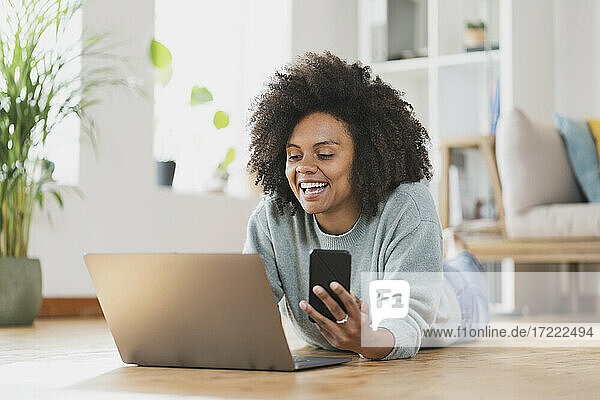 Lächelnde Frau mit Mobiltelefon und Laptop  während sie zu Hause auf dem Boden liegt