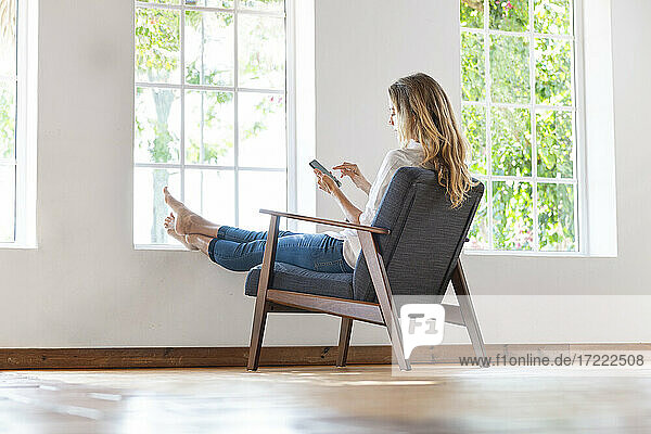 Entspannte Frau  die auf einem Stuhl zu Hause ihr Smartphone benutzt