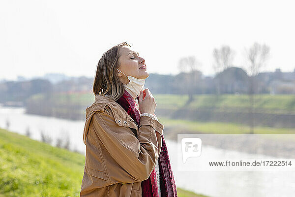 Smiling woman removing face mask while standing by riverbank