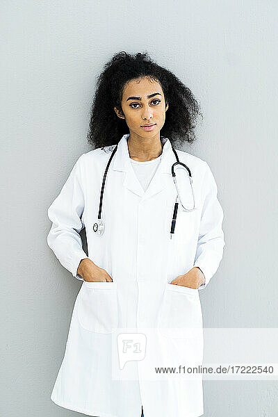 Young female doctor with hands in pockets standing in front of wall