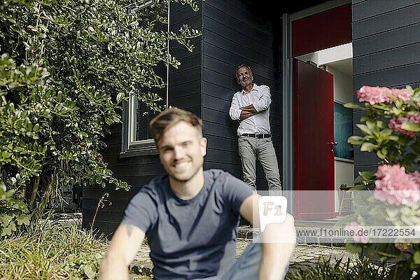 Lächelnder Vater lehnt sich an die Hauswand  während sein Sohn im Hinterhof sitzt