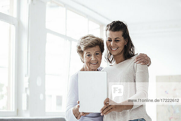 Lächelnde junge Frau mit Großmutter nimmt Selfie durch digitale Tablette zu Hause