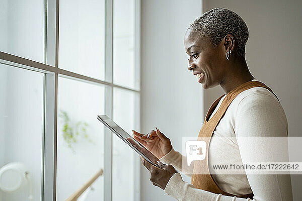 Lächelnde Frau im mittleren Erwachsenenalter  die ein digitales Tablet am Fenster im Wohnzimmer benutzt