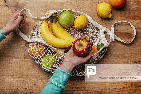 Wiederverwendbare Baumwoll-Netztasche mit frischen Früchten und der Hand eines Mannes  der einen reifen Apfel hält