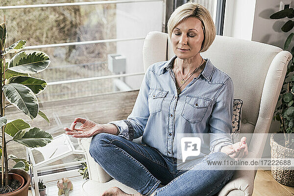 Woman with eyes closed sitting in lotus position on armchair at home