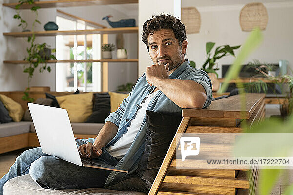 Thoughtful male freelance worker looking away while sitting with laptop on sofa
