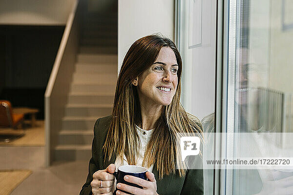Nachdenkliche Geschäftsfrau mit Kaffeetasse  die durch ein Fenster im Büro schaut