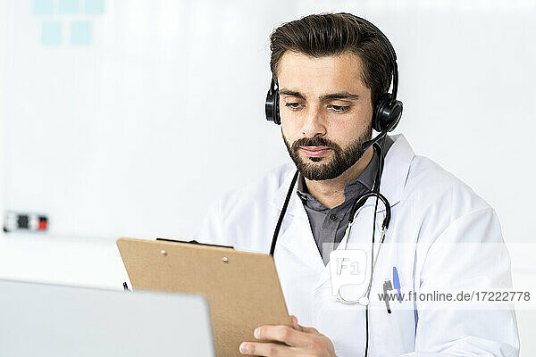 Hübscher männlicher Arzt trägt Kopfhörer  während er einen Bericht auf einem Klemmbrett im Krankenhaus liest