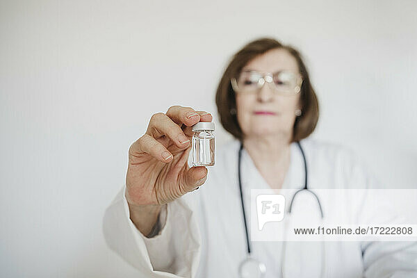 Ältere Ärztin hält Fläschchen vor einer weißen Wand