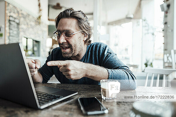 Lächelnder Mann gestikuliert während eines Videoanrufs über einen Laptop in einem Café