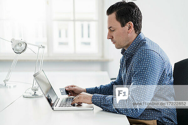Male professional using laptop while holding credit card at workplace
