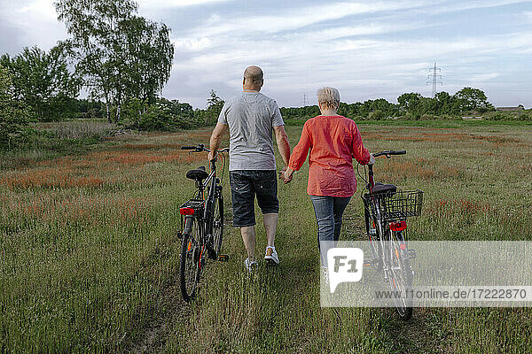 Älteres Paar hält sich an den Händen  während es mit dem Fahrrad auf einem Feld fährt