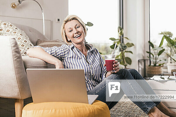 Happy mature woman holding juice glass while watching on laptop at home