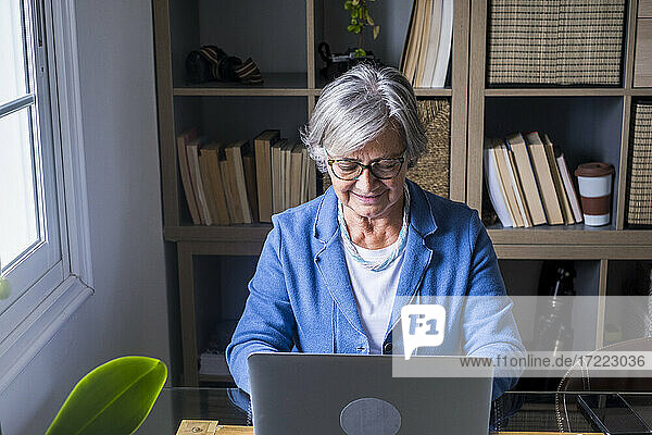 Lächelnde Unternehmerin bei der Arbeit am Laptop vor dem Bücherregal zu Hause