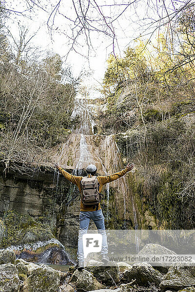 Mittlerer erwachsener Mann steht mit ausgestreckten Armen vor einem Wasserfall im Wald