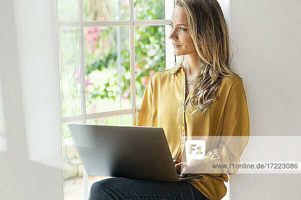 Nachdenkliche Frau  die wegschaut  während sie einen Laptop am Fenster zu Hause benutzt