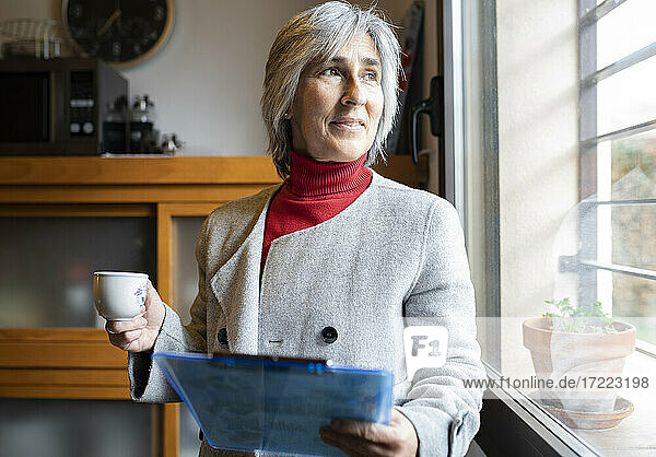 Frau mit weißen Haaren trinkt Kaffee und hält ein Klemmbrett am Fenster zu Hause