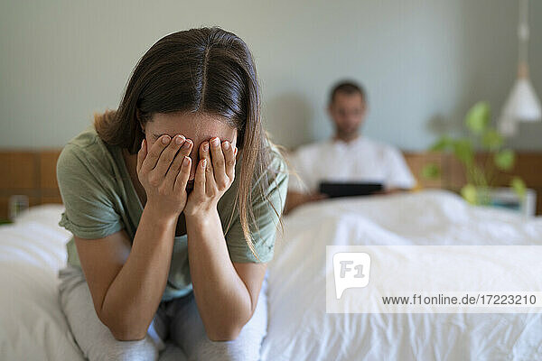 Verzweifelte Frau  die ihr Gesicht bedeckt und auf dem Bett sitzt  während ihr Freund im Hintergrund ein digitales Tablet benutzt