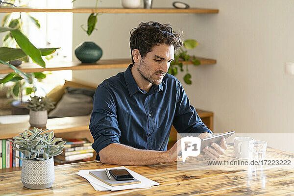 Nachdenklicher männlicher Freiberufler  der ein digitales Tablet benutzt  während er am Tisch im Heimbüro sitzt
