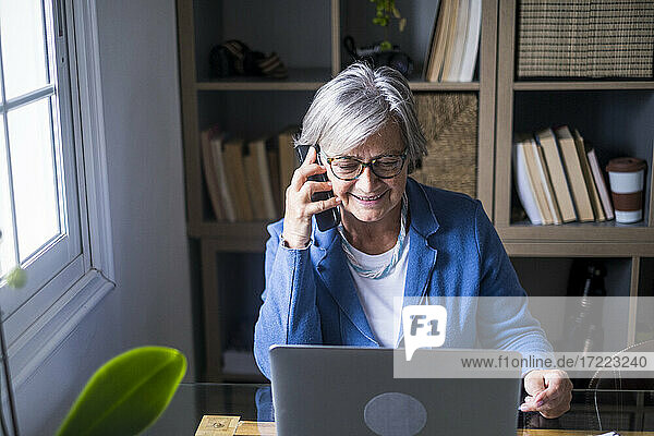 Lächelnde ältere weibliche Fachkraft  die mit einem Mobiltelefon spricht  während sie auf einen Laptop im Heimbüro schaut