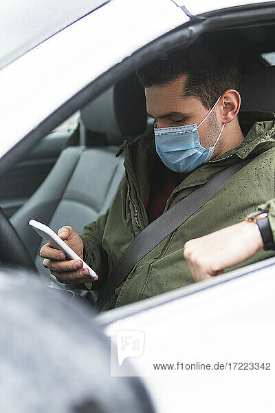 Mann mit Gesichtsschutzmaske  der im Auto sitzend ein Mobiltelefon benutzt