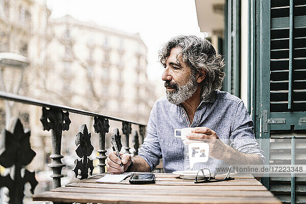 Älterer Mann  der Kaffee trinkt und in einen Notizblock schreibt  während er auf dem Balkon eines Hauses wegschaut