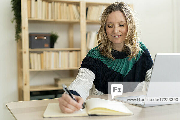 Smiling woman writing in book while sitting by laptop at home