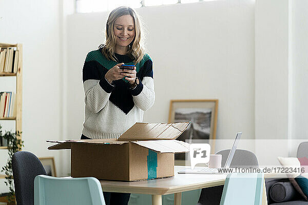 Lächelnde Frau  die ein Foto von einem Paketkarton durch ein Mobiltelefon zu Hause macht