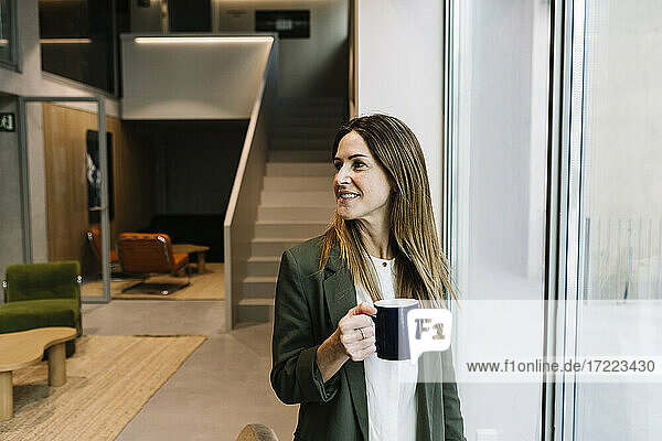 Geschäftsfrau  die wegschaut und einen Kaffeebecher hält  während sie in der Nähe des Fensters im Büro steht