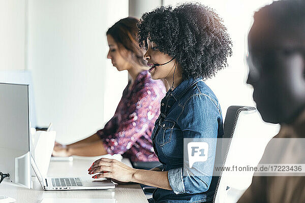 Weibliche Kundenbetreuerin  die einen Laptop benutzt  während sie inmitten von Kollegen im Büro sitzt