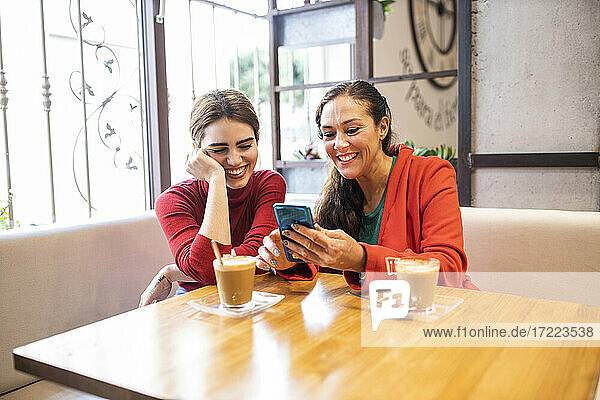 Fröhliche Frau zeigt einer Freundin in der Cafeteria ihr Mobiltelefon