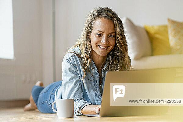 Lächelnde Frau auf Hartholzboden liegend  während sie einen Laptop zu Hause benutzt