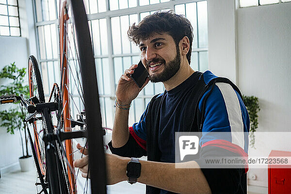 Smiling young man talking on smart phone while holding bicycle wheel spokes at home