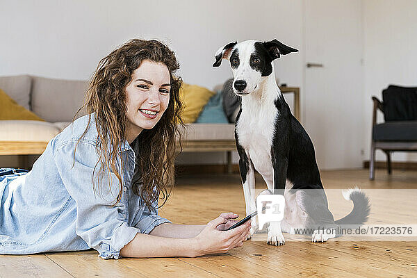 Hübsche Frau liegt auf dem Holzboden und benutzt ein Smartphone  während der Hund neben ihr sitzt und beide in die Kamera schauen