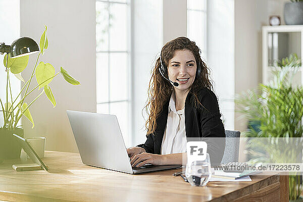Lächelnder weiblicher Kundenbetreuer mit Kopfhörern  der wegschaut  während er vor einem Laptop am Schreibtisch sitzt